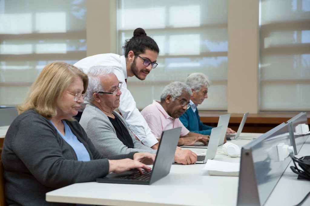 a-man-teach-a-group-of-elderly-to-use-a-laptop-7983552 Senior adults use laptops in a classroom setting, guided by a young instructor.