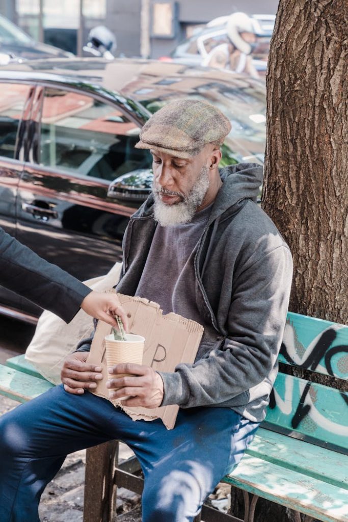 photo-of-a-beggar-receiving-money-from-a-person-9532336 An elderly man seated on a bench receives money in an urban setting.